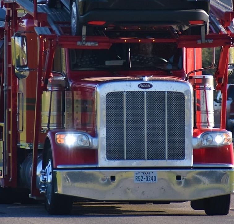 The chrome grille and lit headlamps of a conventional truck pulling a loaded car carrier.