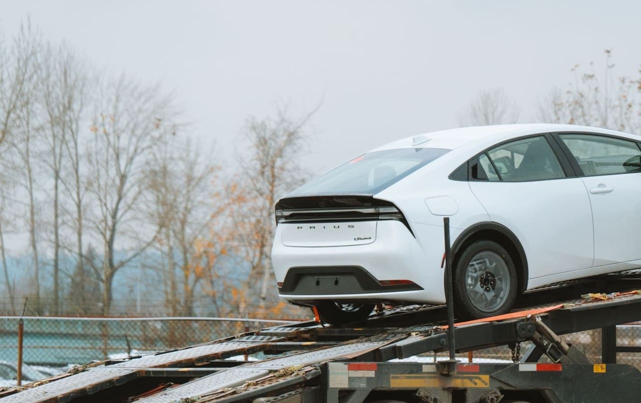 A new sedan standing on the angled deck of a car carrier, its wheels held by a ratchet strap over the steel grating.